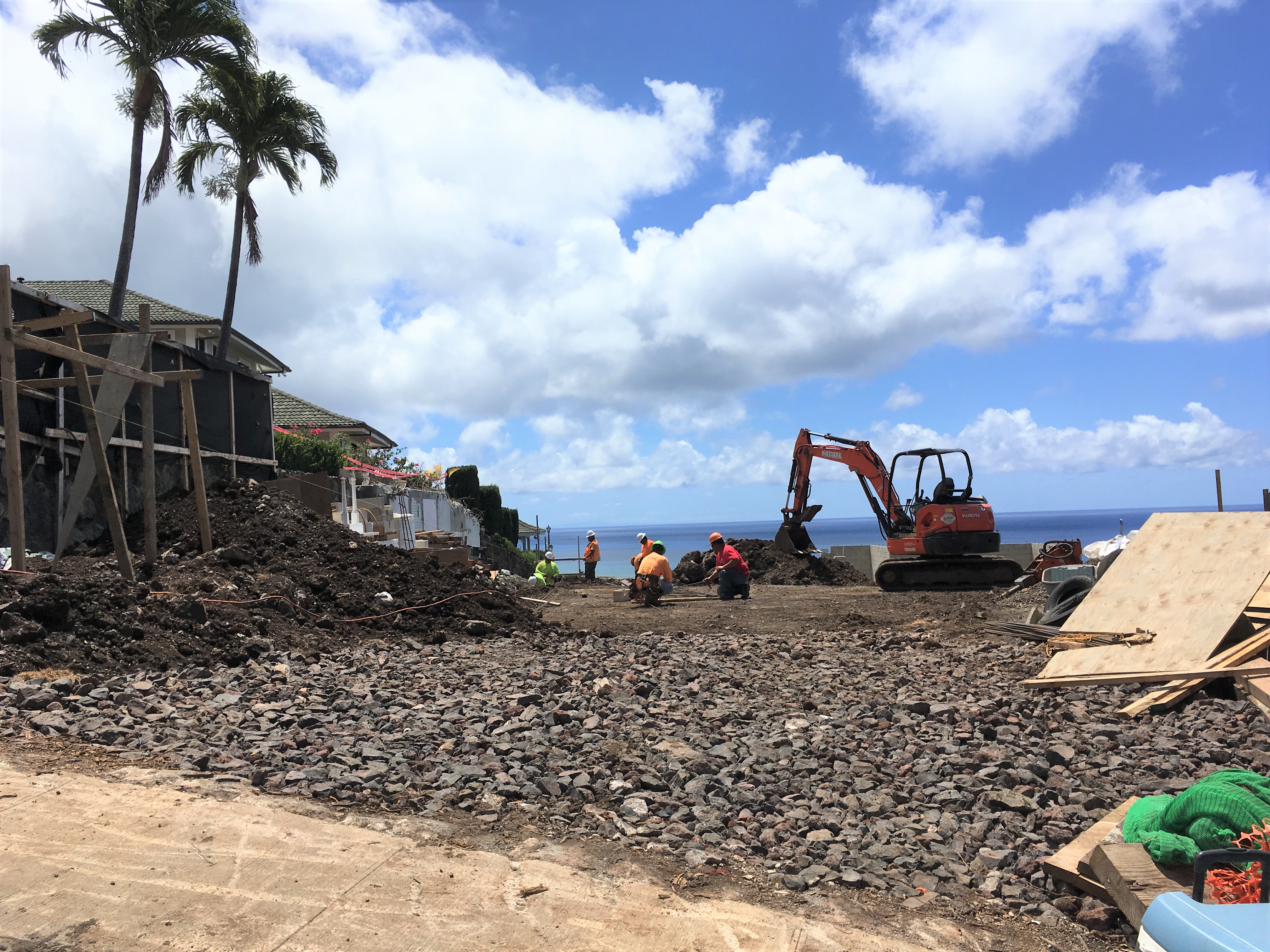 construction, street view, sidewalk and rocks to trap debris in foreground, earth mover and construction workers further back, with ocean and cloudy blue sky in distance