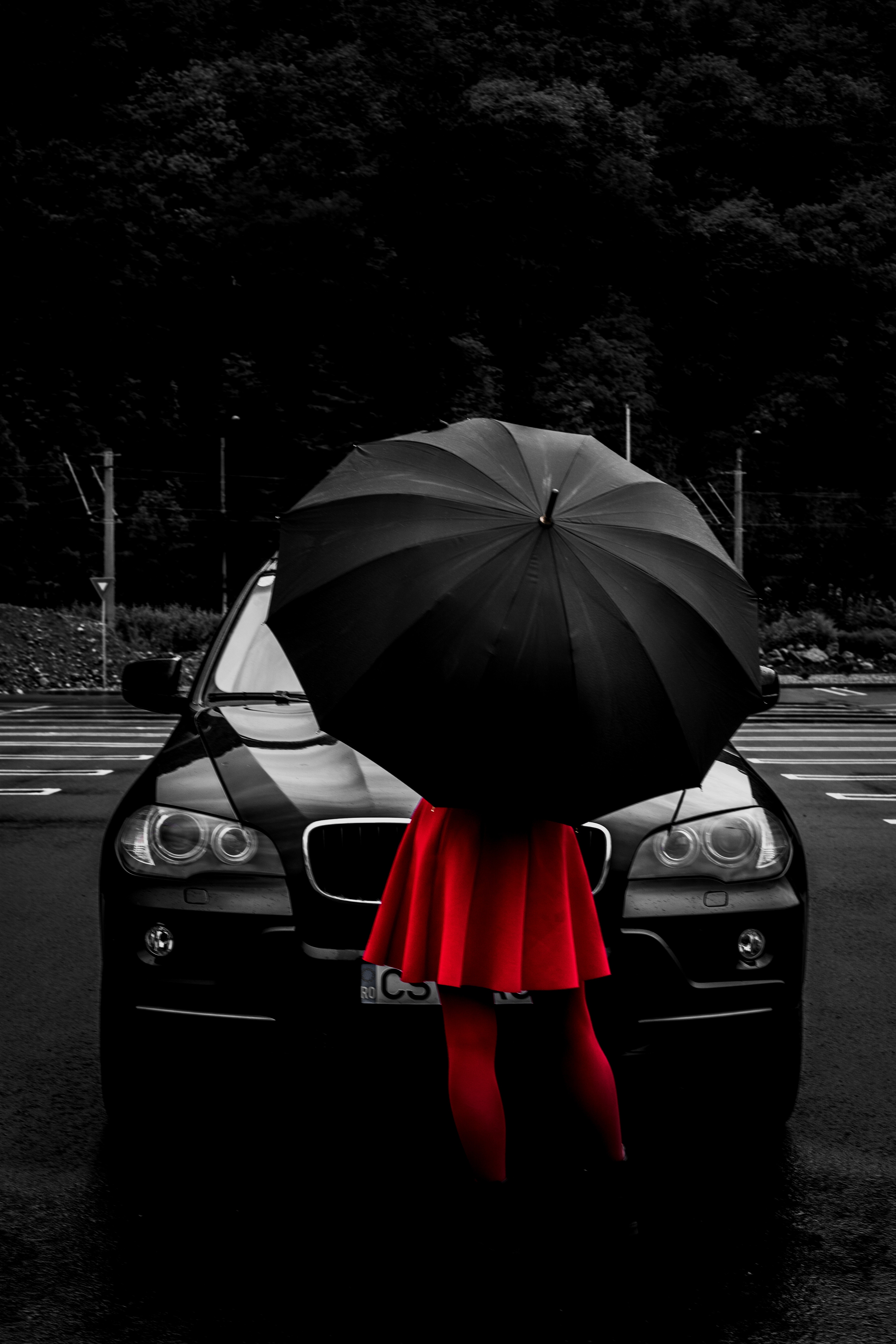 black car facing camera with female in red dress and red tights holding black umbrella standing facing car