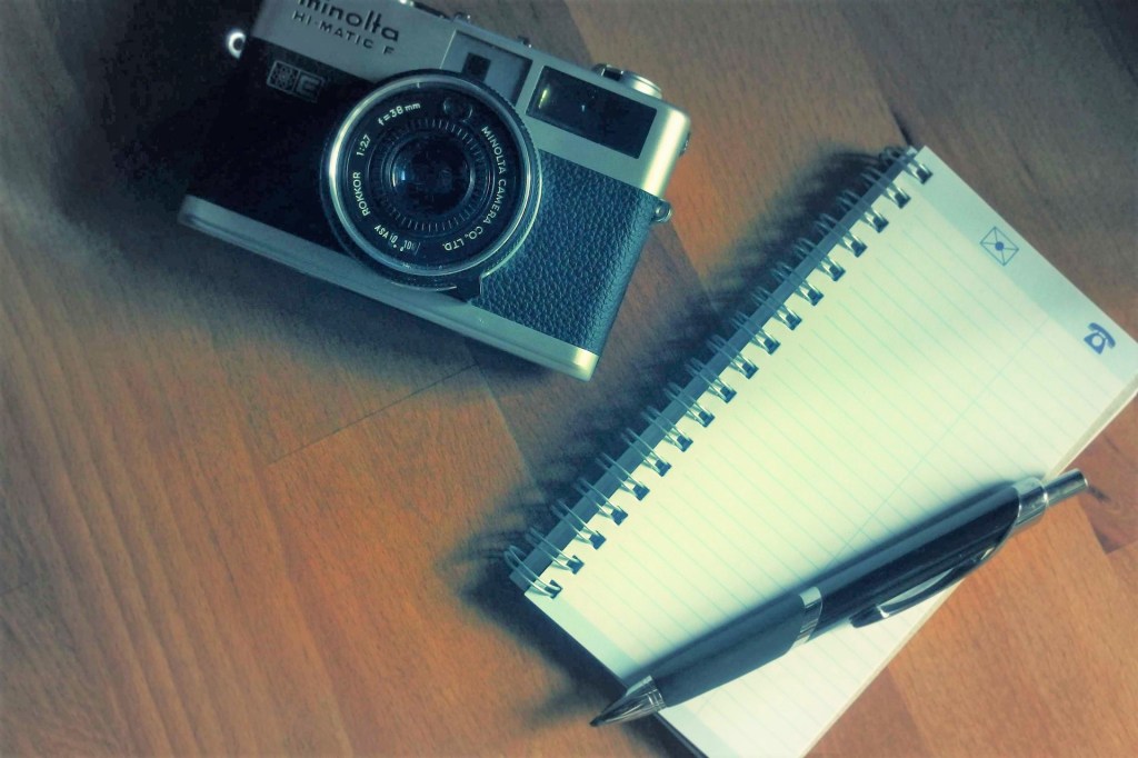 wooden surface with old-fashioned camera next to small notebook with ballpoint pen on top