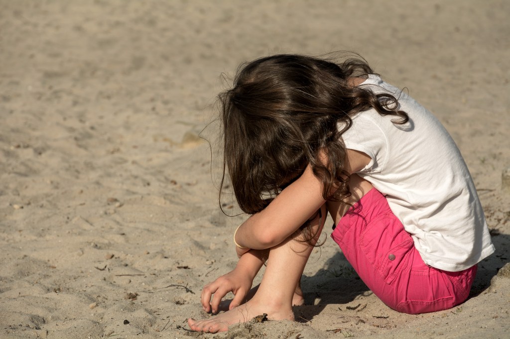 brown-haried girl in white t-shirt and dark pink shorts sitting on beach with head on knees