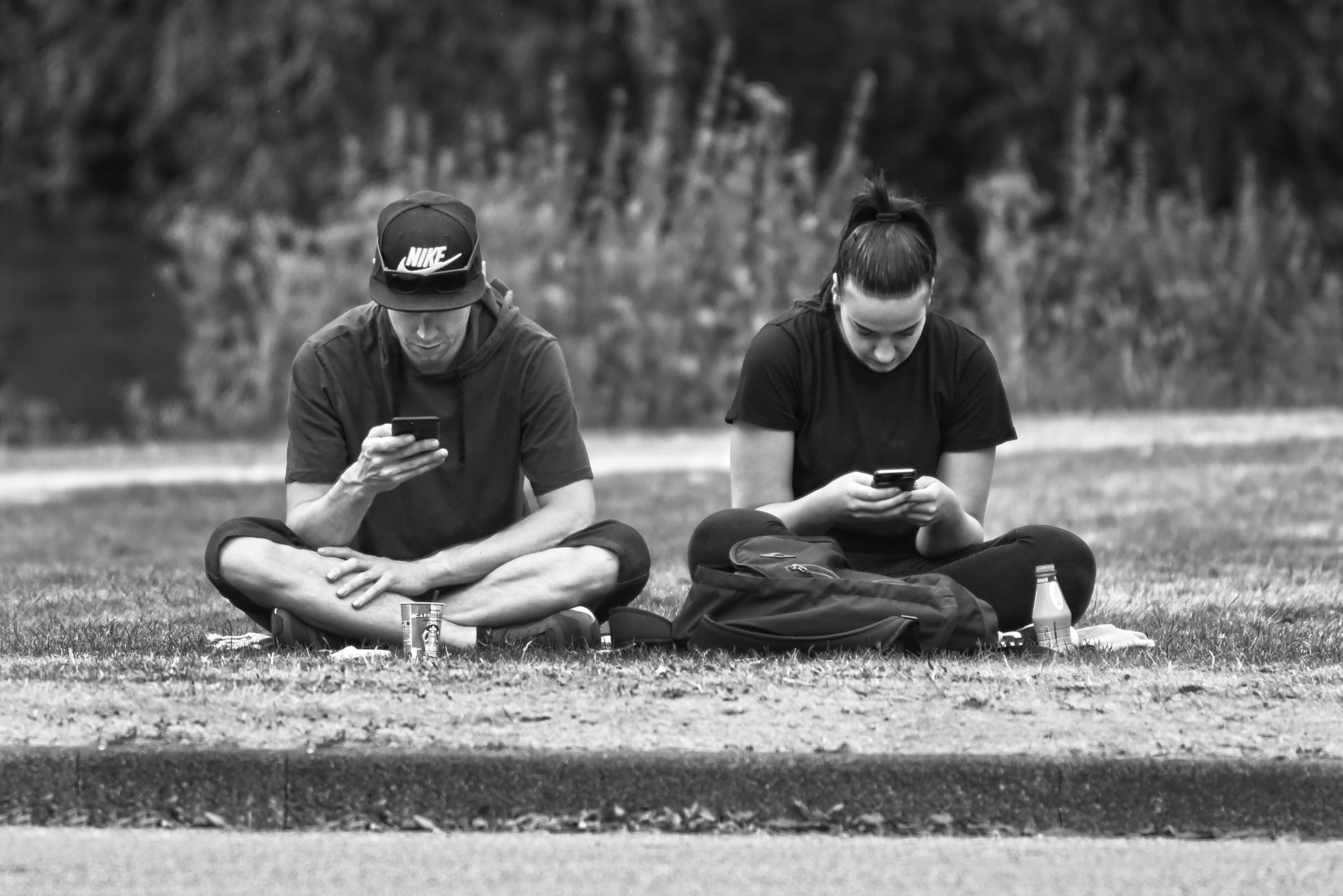 black and white photo of man and woman sitting next to each other in park while using their phones