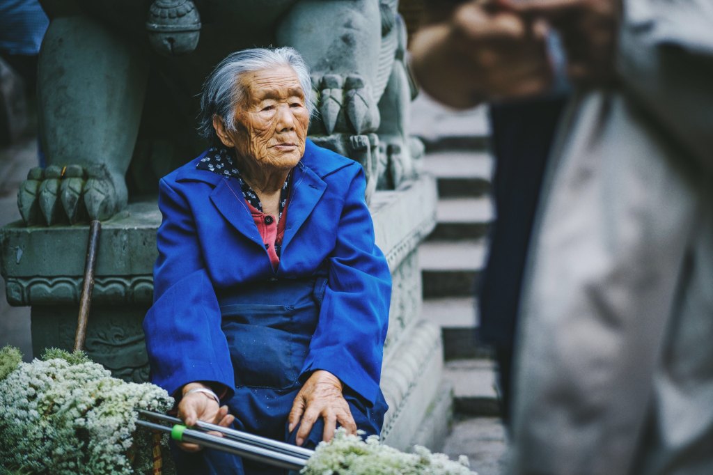 old Asian woman in blue coat sitting in front of stone statue