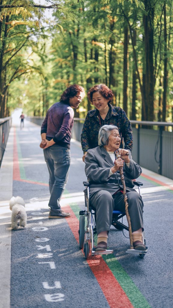 three Asian women and a small dog on a pedestiran path, one is pushing an elderly woman in a wheelchair